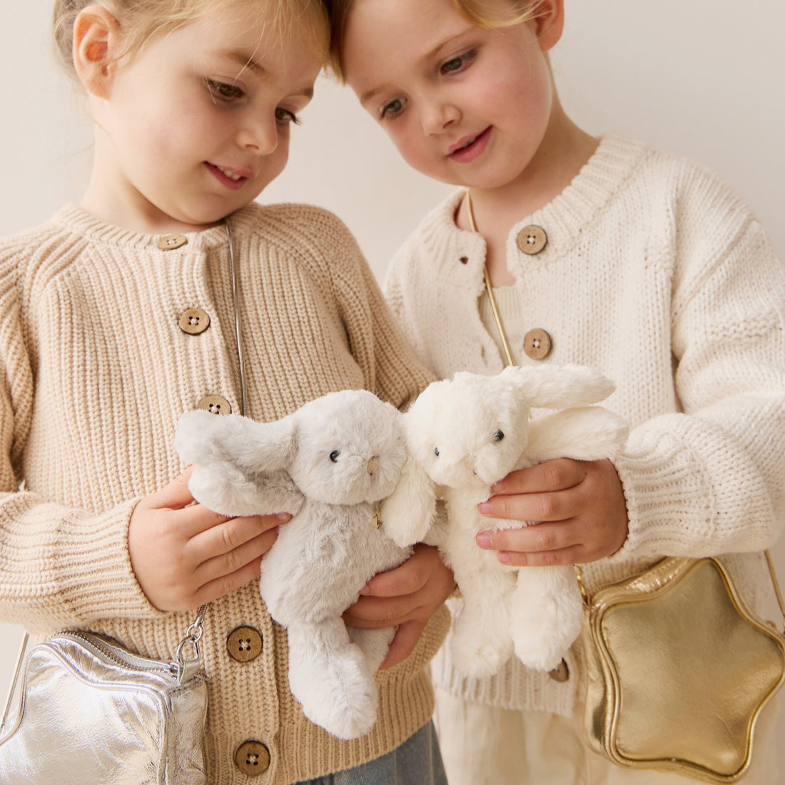 Two young girls in sweaters, holding stuffed bunnies, and wearing small crossbody bags.