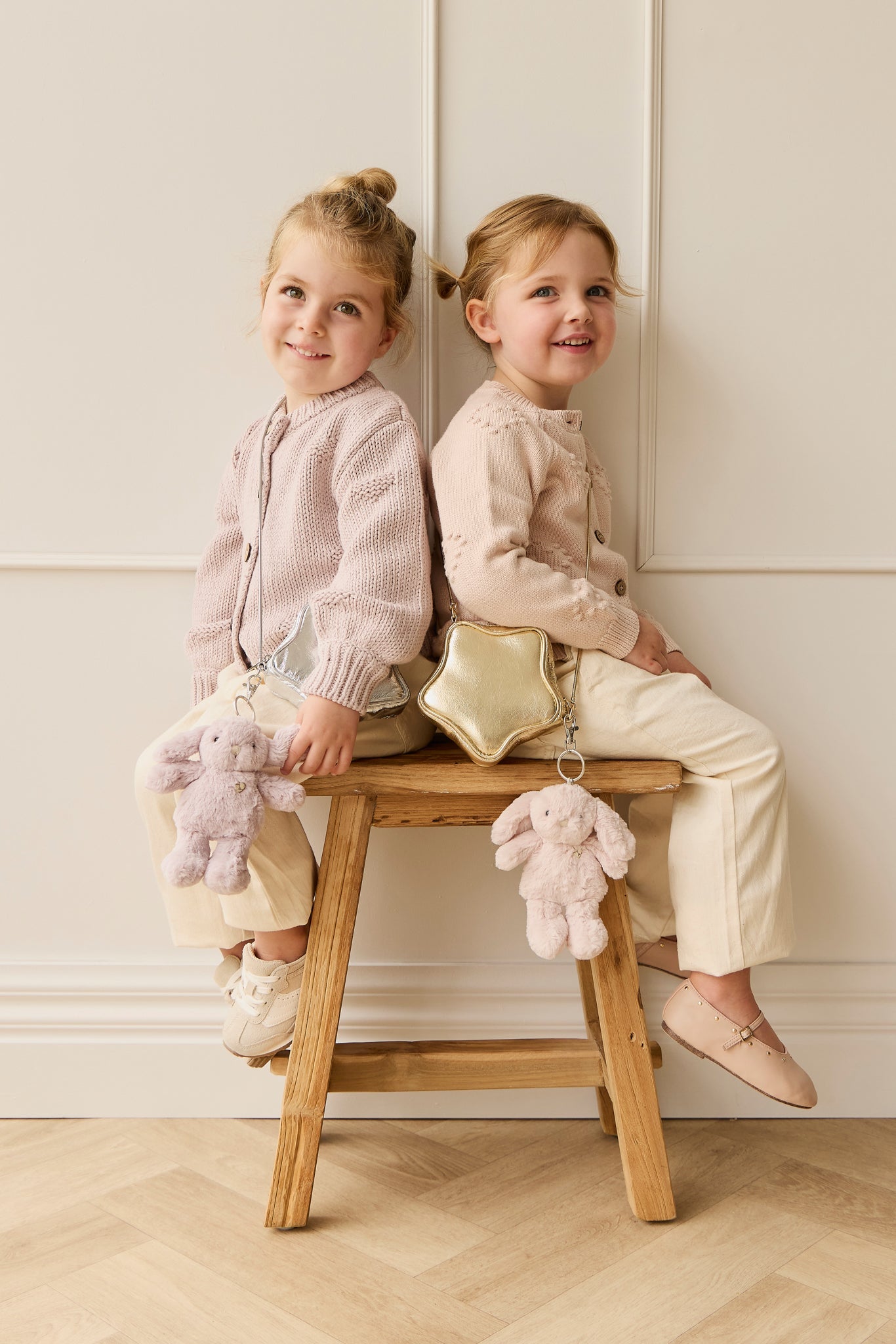 Two young girls in pink sweaters and cream pants sit back-to-back on a wooden stool with a gold star-shaped purse and stuffed bunny.