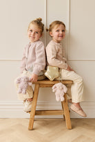 Two young girls in pink sweaters and cream pants sit back-to-back on a wooden stool with a gold star-shaped purse and stuffed bunny.