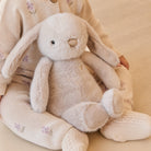 Soft beige plush bunny toy with long floppy ears and black button eyes, held by a child in floral knit outfit, front view