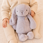 Soft gray plush bunny with long floppy ears and golden heart tag, held by child in cream knit outfit, front view.