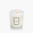Lit candle in a decorative glass container with 'Voluspa' branding on a white background