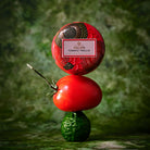 A stacked product display featuring a red round decorative container labeled 'VOLUSPA TOMATO TRELLIS' on top of a fresh red tomato, which is balanced on a green cucumber, set against a textured green background.