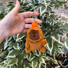 A plush bear keychain with an orange on its head held against a leafy background.

