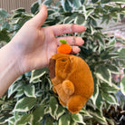 A hand holding a brown, furry, stuffed capybara keychain with an orange on its head.
