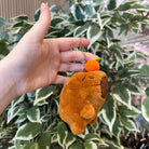 A hand holds a brown, plush capybara keychain with a small orange fruit on its head, against a background of green and white foliage.
