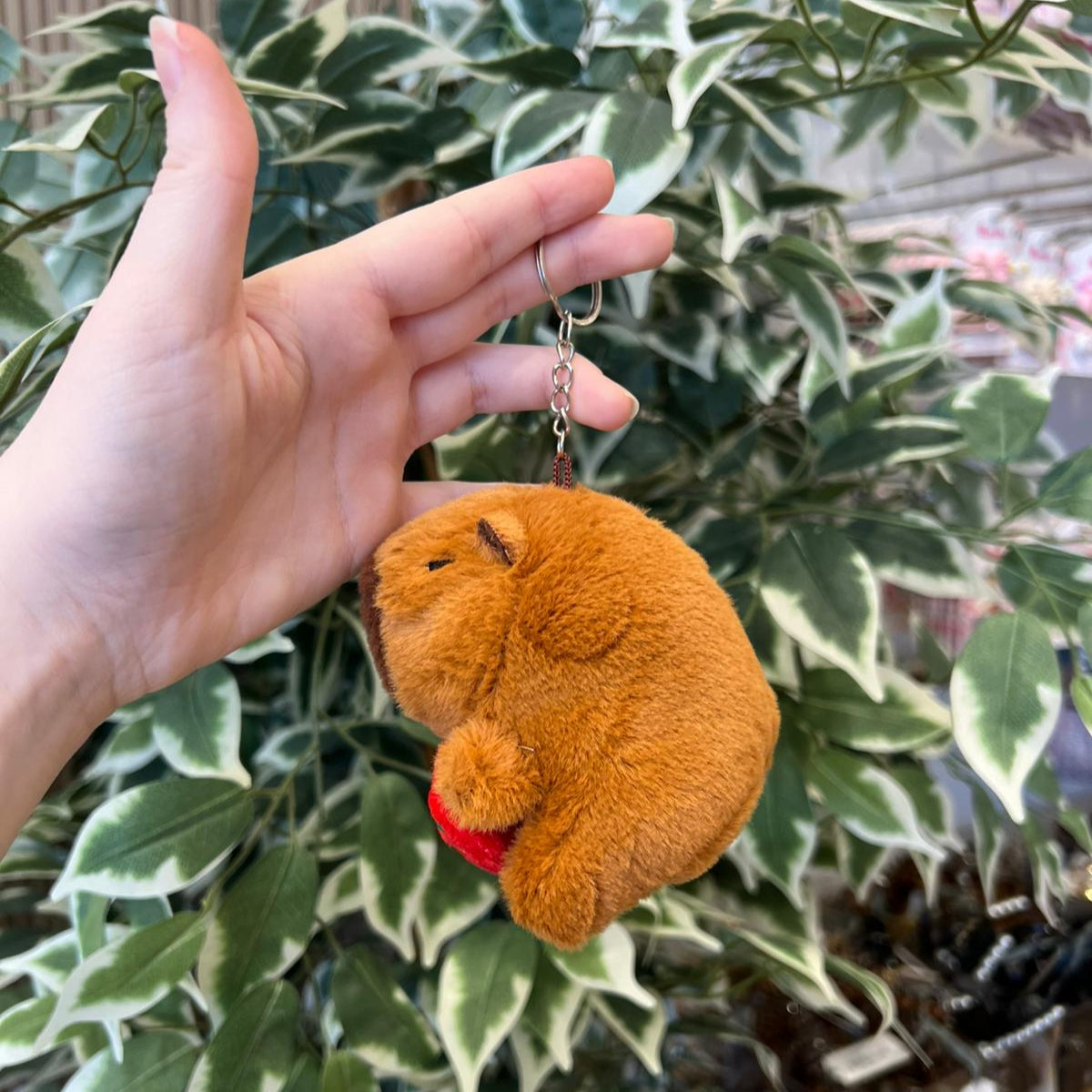 A plush brown capybara keychain being held against a leafy plant.
