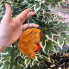 A hand holds a capybara plush keychain with a red heart, against a leafy green backdrop.