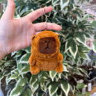 A brown, stuffed bear keychain is being held in front of a plant.