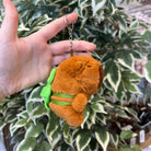 A hand holds a brown plush capybara keychain with a green leaf detail against a background of green and white leaves.
