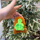 A hand holding a keychain with a plush turtle on top of a plush bear in front of a plant.