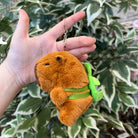 A hand holds a plush, orange capybara keychain with a green plant tied to its back, set against a background of green and white variegated leaves.