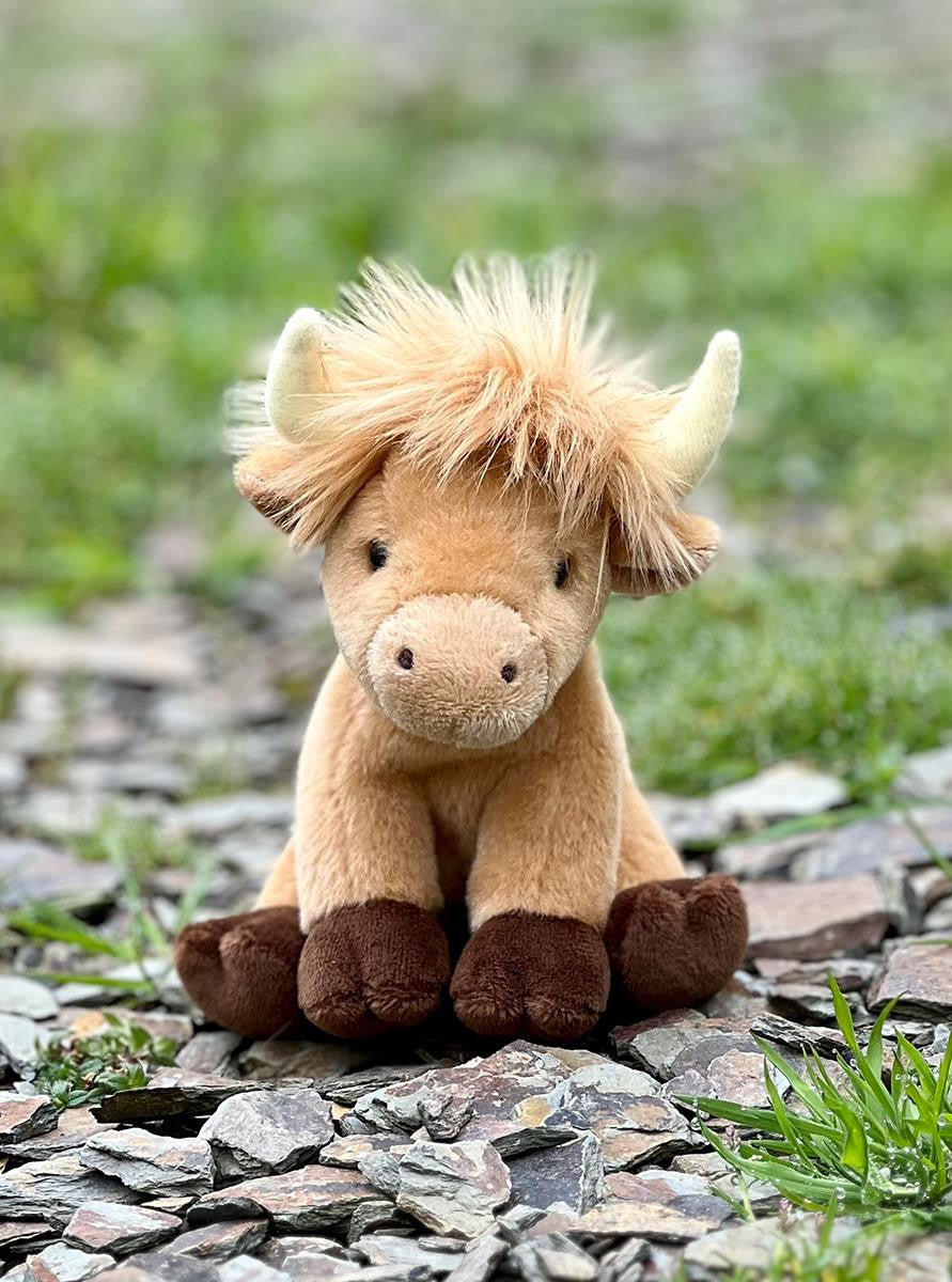 Plush toy Highland cow sitting on a stone path with grass in the background