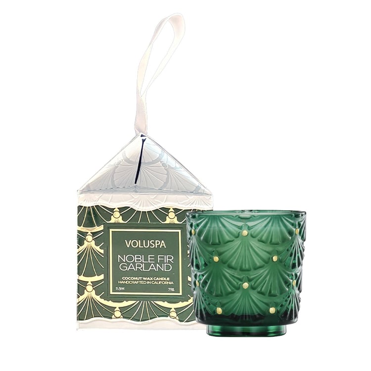 Voluspa Noble Fir Garland coconut wax candle in a green textured glass holder with a matching patterned box and white hanging ribbon, shown on a white background.