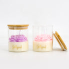 Two clear glass jar candles with bamboo lids on a white background. One candle has a purple flower-shaped wax top, and the other has a pink flower-shaped wax top.