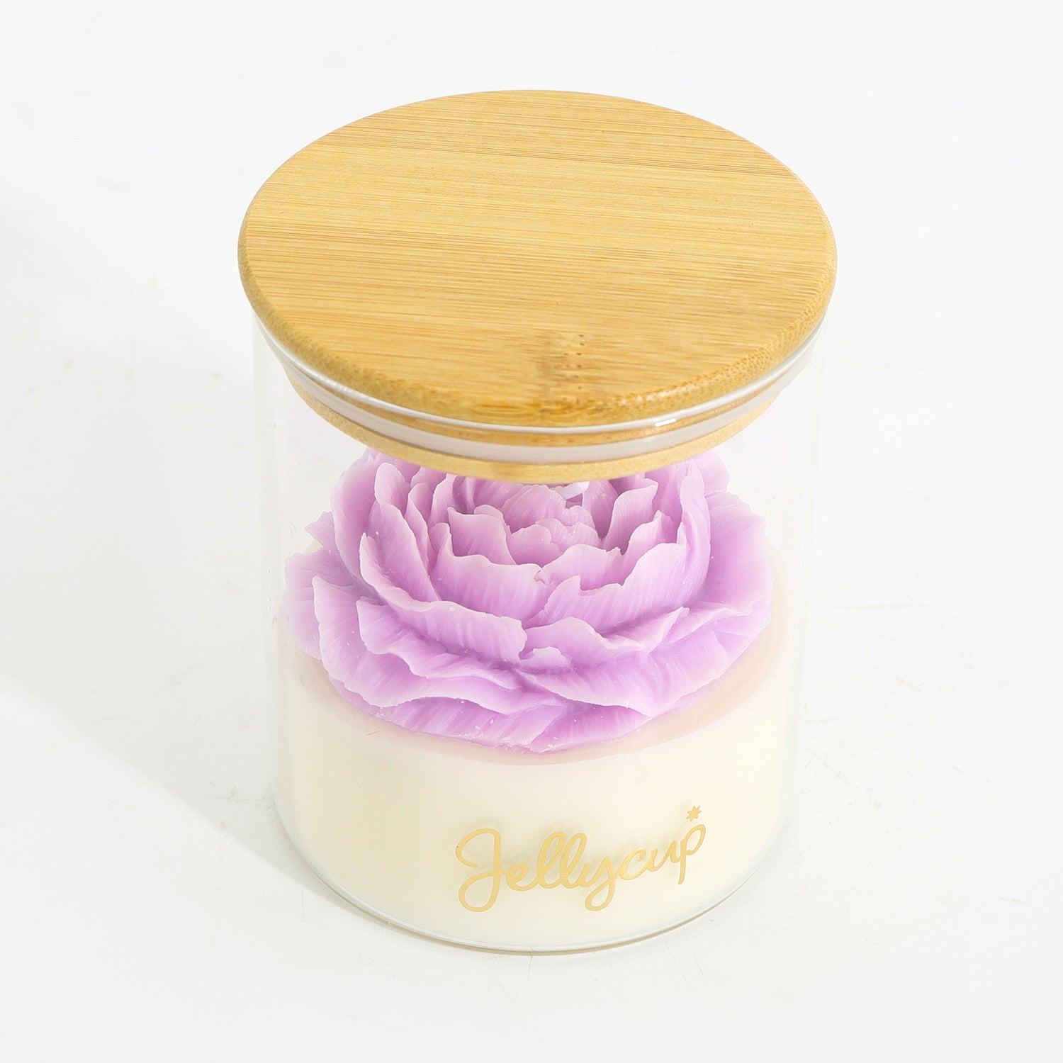 A clear glass jar candle with a light wood lid, featuring a layered design with a pale cream base and a prominent light purple flower on top. The brand name 'Jellycup' is printed in gold script on the front of the jar.