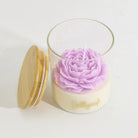 A clear glass jar candle with a light cream wax base, a large lavender-purple flower-shaped wax top, and a gold script logo reading 'Jellycup' on the front. A round bamboo-style lid with a gold rim leans against the jar on a clean white background.