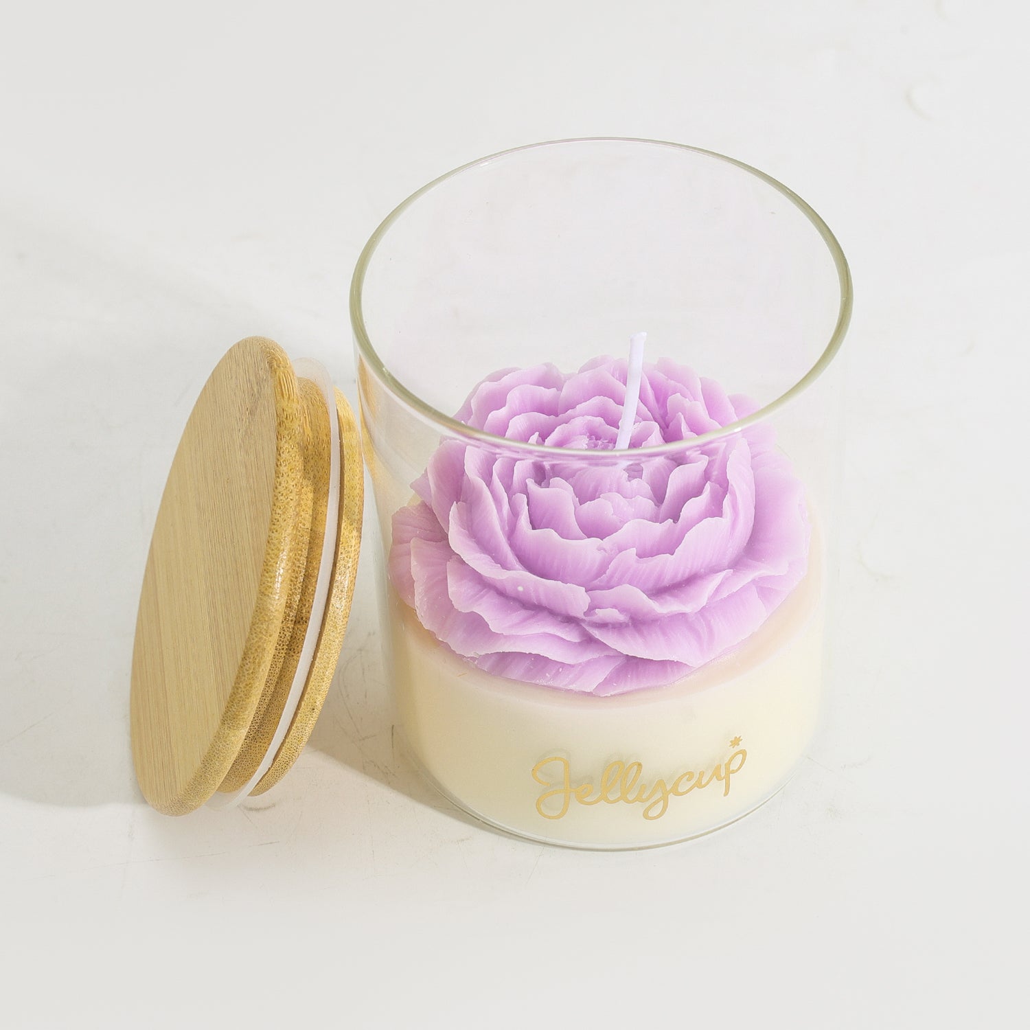 A clear glass jar candle with a light cream wax base, a large lavender-purple flower-shaped wax top, and a gold script logo reading 'Jellycup' on the front. A round bamboo-style lid with a gold rim leans against the jar on a clean white background.