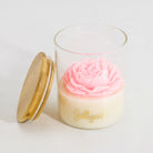A clear glass jar candle with a cream-colored wax base, a pink rose-shaped wax top, and gold foil script reading 'Jellycup' on the front. A light wood lid with a gold rim rests beside the jar on a clean white background.