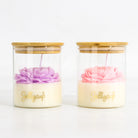 Two glass jar candles with bamboo lids on a white background. The left candle has a purple flower and the right candle has a pink flower, both resting on a cream-colored wax base with gold 'Jellycup' branding.