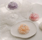 Floral-shaped candle by Jellycup with a decorative paper flag, sitting on a plate near lace fabric.