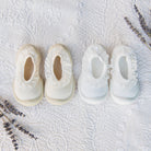 Two pairs of baby slippers, one off-white and one white, with lace trim, on a textured white blanket, accented with sprigs of lavender.
