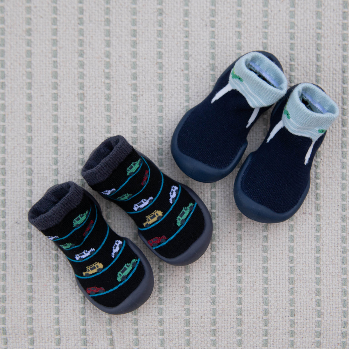 Two pairs of baby socks/shoes with grippy soles on striped woven background. One pair is black with colorful car patterns, the other navy blue with white stripes.