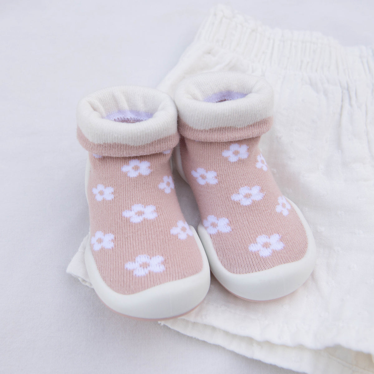 Pair of pink baby shoe-socks with white flowers on white fabric.