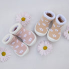 Two pairs of baby shoe-socks with flower patterns and white soles, arranged with pink daisies on a white background.