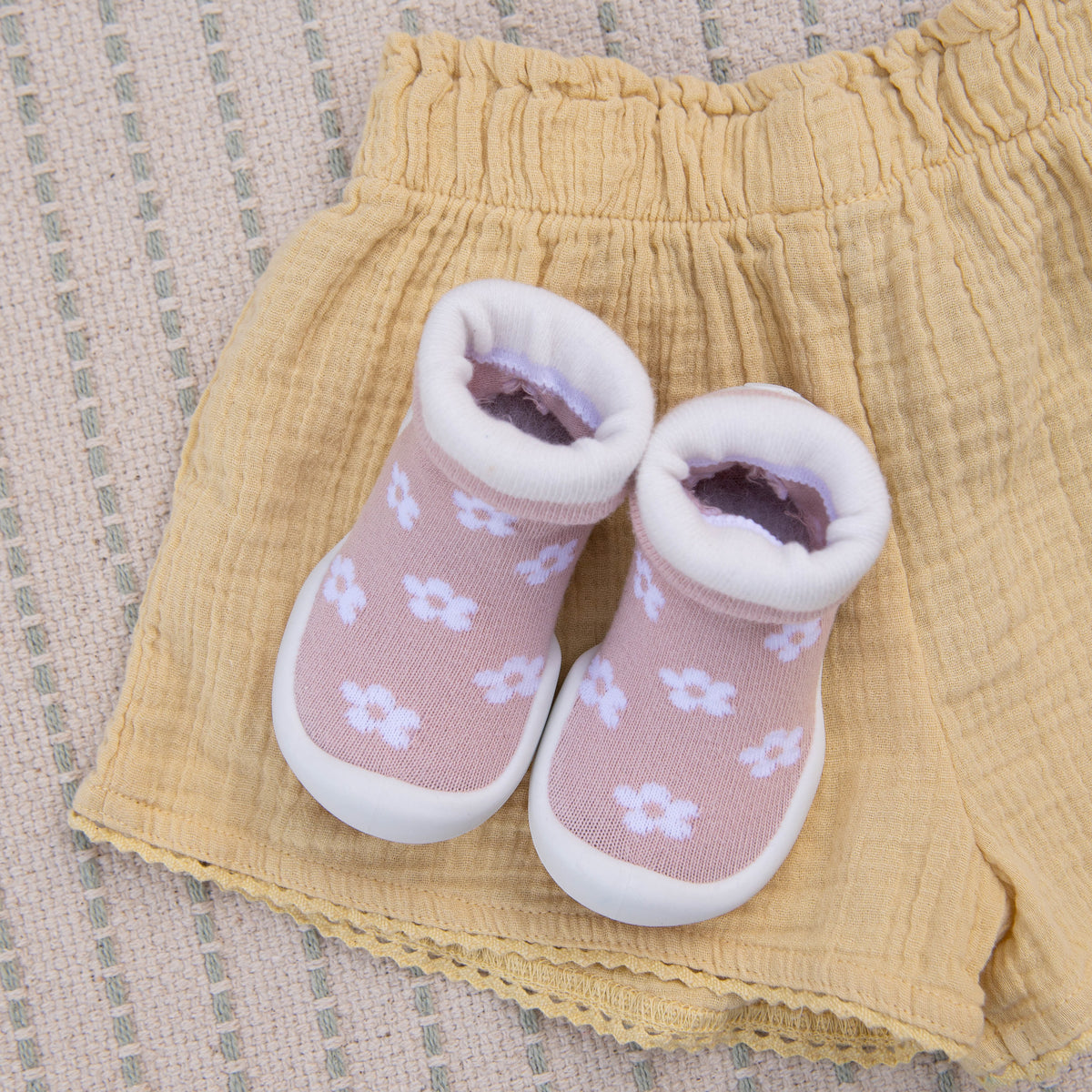 Pair of pink baby shoes with white flower pattern, atop yellow shorts with elastic waist, on a woven textile.
