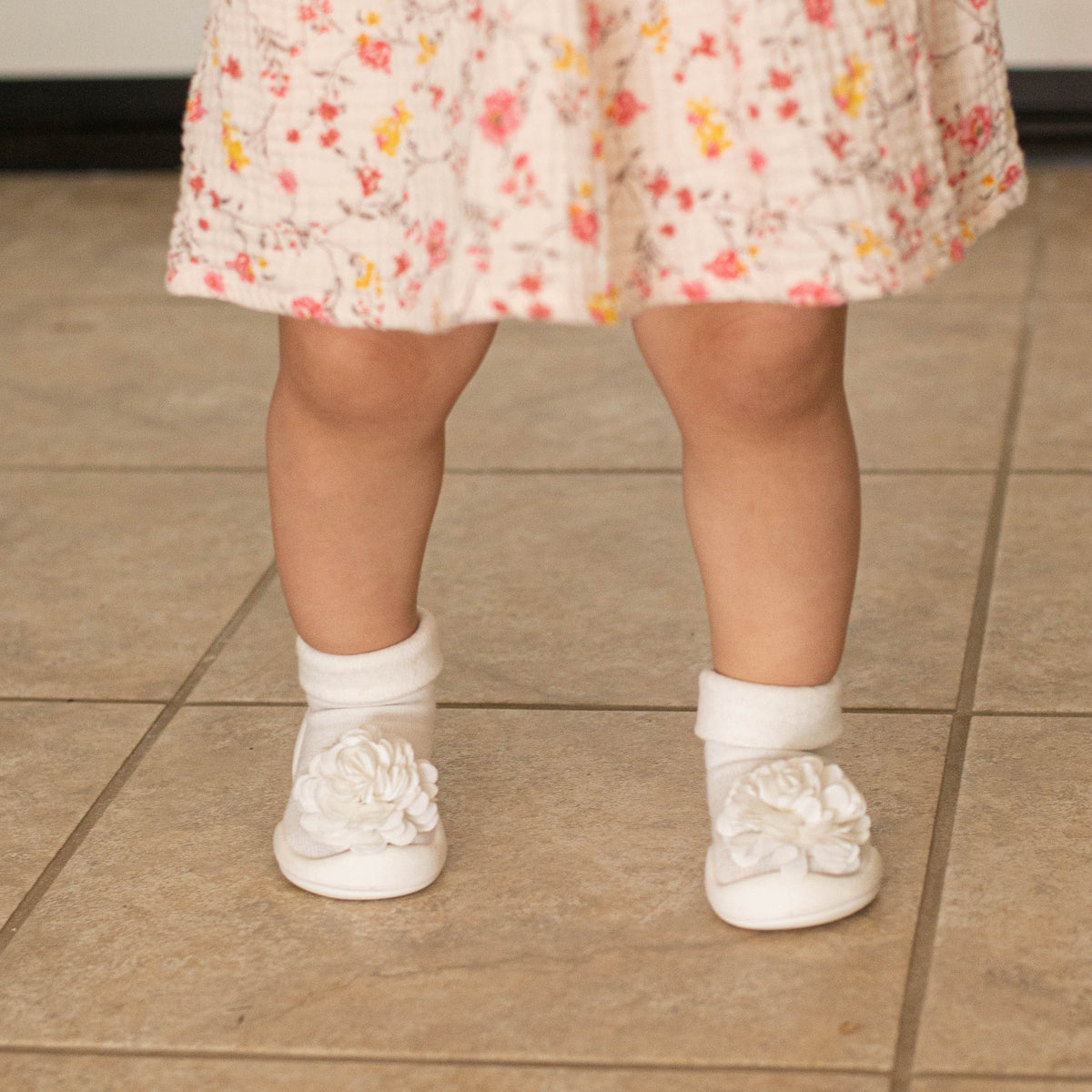 A baby's legs and feet in white socks and shoes, wearing a floral dress, standing on a tile floor.