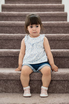 Young child sits on carpeted stairs. Sleeveless floral shirt and denim shorts. Pink and white booties.