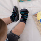 Infant wearing black socks with toy car pattern and gray sock shoes.