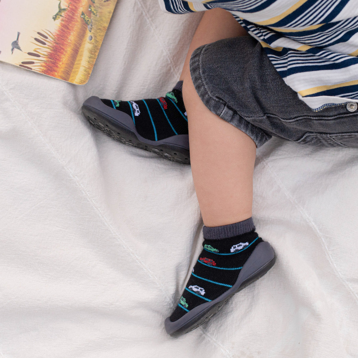 A toddler wearing black socks with colorful cars and gray shoes, seated on a white blanket with a book nearby.
