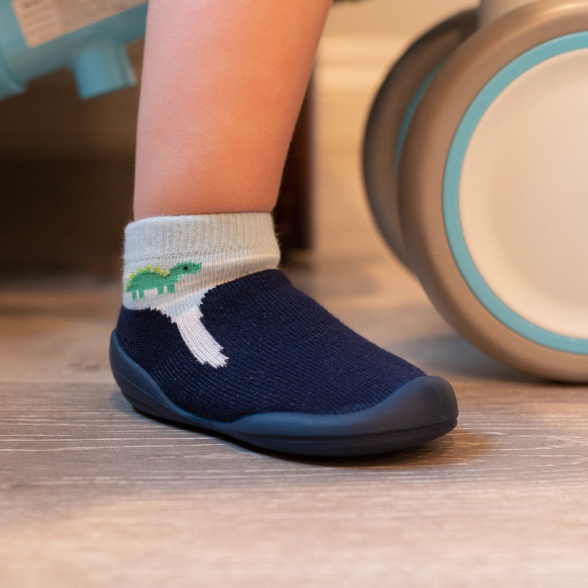 A baby's foot wearing a navy blue sock-shoe with a dinosaur on the sock.
