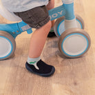 Child on blue balance bike with brown and white wheels; dark-wash shorts; dinosaur socks; and navy shoes.
