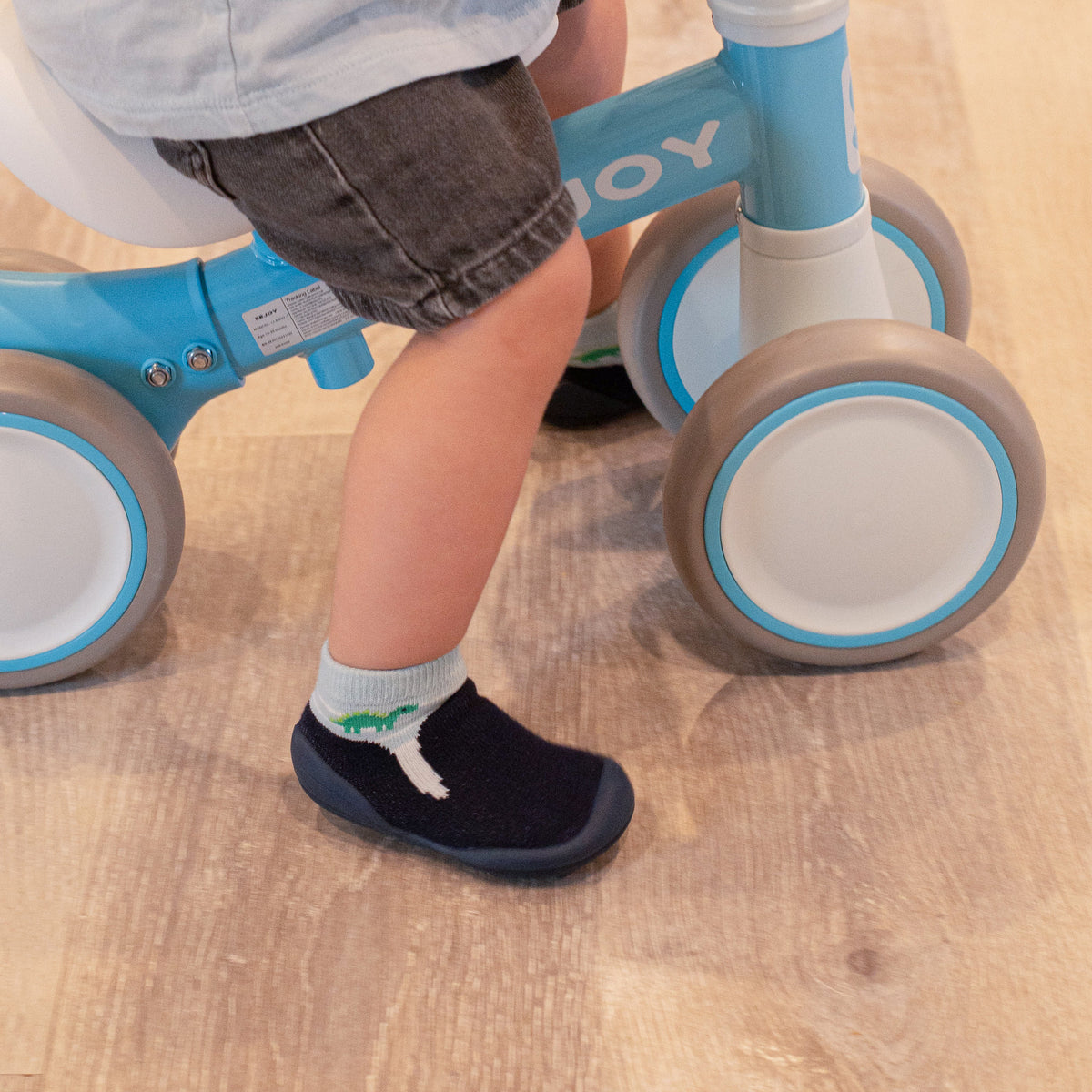 Child on blue balance bike with brown and white wheels; dark-wash shorts; dinosaur socks; and navy shoes.
