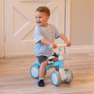 A young child wearing a light blue t-shirt with a seagull graphic, gray shorts, and navy shoes sits on a blue and white balance bike in an indoor setting.