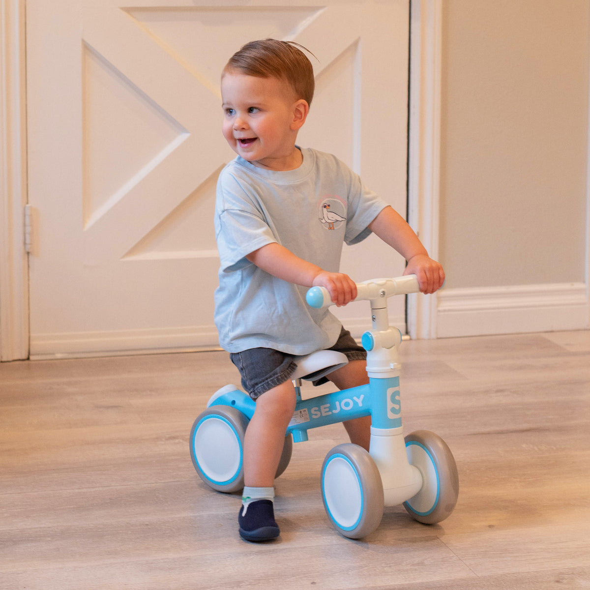 A young child wearing a light blue t-shirt with a seagull graphic, gray shorts, and navy shoes sits on a blue and white balance bike in an indoor setting.