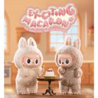 Two furry, rabbit-eared monster dolls stand beside a miniature cake in a kitchen setting. Above them is a logo reading, "Exciting Macaron."