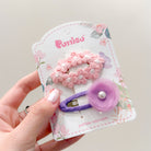 Two flower hair clips attached to decorative packaging. Pink and purple tones.