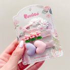 A hand holds a retail display card with three hair clips. The clips feature white daisies, pink flowers, and purple and pink hearts.