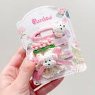 A set of three decorative hair clips with a white, floral-themed cardboard backing. A pink snap clip with a white bear face on top, a pink snap clip with three pink flowers, and a small clip with a white bear face with a pink bow attached to a lacy flower pattern.