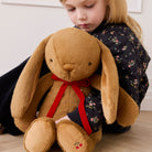 Brown plush bunny with long floppy ears and red velvet bow, by Snuggle Bunnies, held by child in floral jacket, lifestyle view