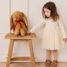 Tan plush bunny with floppy ears and red bow, by Snuggle Bunnies, sitting on wooden stool beside child in floral dress.