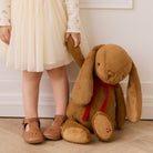 Brown plush bunny toy with long floppy ears and red velvet bow, by Snuggle Bunnies, shown beside child in cream dress and tan shoes, lifestyle view