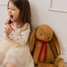 Brown plush bunny with long floppy ears and red velvet bow, by Snuggle Bunnies, sitting beside laughing child in floral dress, lifestyle side view