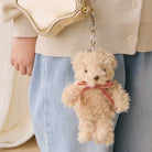 Plush beige teddy bear keychain with red striped bow, attached to cream bag, worn by person in beige sweater and blue jeans, front view