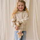 Plush beige bear keychain with red striped bow, attached to gold pouch, held by child in cream cardigan and jeans, lifestyle front view