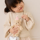 Plush tan bear keychain with pink party hat and bow, by Snuggle Bunnies, held by child in cream sweater, lifestyle front view